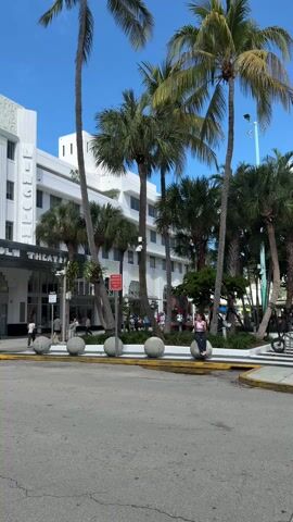 The camera faces an intersection on a sunny day in Miami, Florida, across from the Art Deco styled Lincoln Theatre.  Palm trees line the street and sidewalk in front of the theater. A person sits on one of the large concrete spheres lining the curb.  People walk on the sidewalk.  Traffic passes on the street.  The sky is blue.