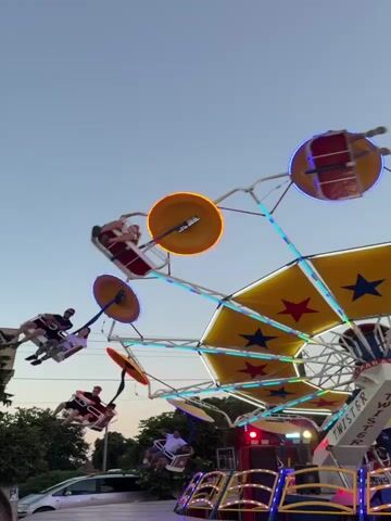 This video captures a thrilling amusement park ride at dusk in Koinare, Bulgaria. The ride, identified by visible text as "TWISTER," is a large, spinning attraction with multiple swinging gondolas. The main subject is the ride itself, a complex structure of metal beams adorned with colorful lights and a large, star-patterned yellow canopy. Several people are visible in the gondolas, their bodies angled outwards as the ride spins and swings them through the air. They appear to be enjoying the exhilarating experience.

The time of day is evident from the soft, fading light and the clear, pale blue sky. The weather appears to be fair, with no signs of rain or wind. The setting includes some trees and a hint of buildings in the background, suggesting a location on the outskirts of a town or within a larger park area. A silver car is parked in the lower left corner, adding a touch of everyday reality to the scene. The overall mood is one of fun and excitement, typical of an amusement park environment. The lights on the ride are a prominent feature, pulsing and changing color, adding to the festive atmosphere. The visible text "TWISTER" on the ride's central structure clearly identifies the specific attraction.