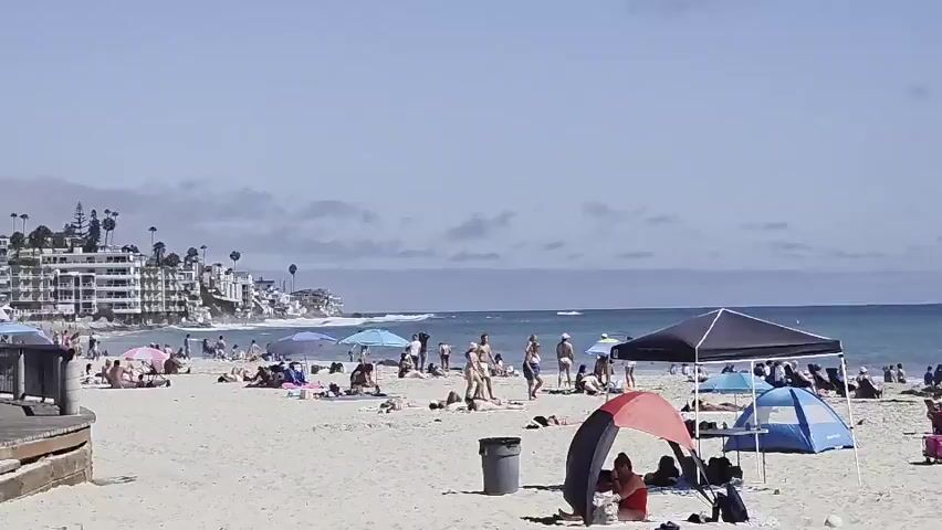 The video displays a crowded beach scene on a sunny day with a clear blue sky and scattered clouds. The ocean is visible in the background with moderate waves. On the sandy beach, numerous people are relaxing, sunbathing, and walking. Several beach umbrellas and pop-up tents provide shade. In the foreground, a black pop-up canopy is set up, with a red and black beach umbrella to its left. A blue tent is positioned near the black canopy. Further to the right, a white lifeguard tower stands prominently on the sand. People are scattered across the beach, some sitting in chairs or on towels, while others are strolling or playing near the water. A trash can is visible near the foreground. The overall atmosphere is one of a typical busy summer day at the beach.