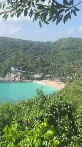 The video shows a scenic view of a tropical beach from a high vantage point. Lush green vegetation frames the shot in the foreground and along the sides, partially obscuring the view. The beach, with tan-colored sand, curves along the edge of a bay of clear, turquoise water. Buildings are nestled along the beach and on rocky outcrops to the left of the frame. A boat is anchored in the water. The background features a densely forested hillside under a sky with scattered clouds. A tall white structure resembling a communications tower can be seen atop the hill. The overall scene depicts a serene and tropical paradise.
