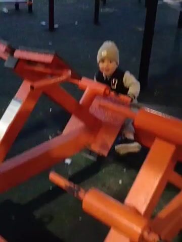 A young child wearing a light brown hat and a jacket with a white collar and a black number "9" on the chest is sitting on an orange metal exercise machine. The child is facing away from the camera, looking at the machine. The machine appears to be a leg press or similar weight training equipment. The child is in a playground or outdoor gym setting, indicated by the dark ground surface and other exercise equipment visible in the background. The lighting suggests it may be dusk or evening. The child's hands are on the handles of the machine. The machine has various orange metal bars and levers.