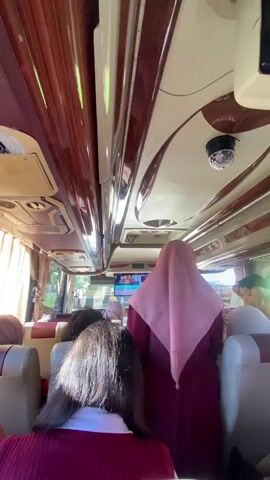 A video shot from a low angle inside a bus shows a group of children and adults. The camera pans from left to right, revealing the bus interior with beige and brown accents. Several individuals wearing hijabs are visible. A woman in a pink hijab and maroon dress stands in the aisle, talking and gesturing. Other children and adults are seated, some looking at the camera. A small disco ball light hangs from the ceiling. A monitor displaying a video is mounted on the front ceiling. The bus windows show a bright exterior, possibly indicating the bus is parked or moving slowly.