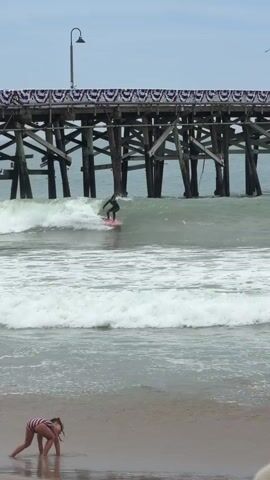 A surfer rides a wave in the ocean near a wooden pier. A woman with long brown hair stands on the beach holding a football. In the foreground, a child in a striped swimsuit crawls on the sand. The sky is cloudy.
