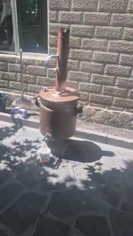 The video shows a large, rustic, copper-colored distillation apparatus, possibly a still or boiler, sitting outside on a paved area next to a brick wall. The apparatus is composed of a large cylindrical body with a smaller, narrower pipe extending vertically from the top. The main body has a spigot at the bottom, under which sits a small, partially filled bucket. A window is visible to the left of the apparatus along with a bucket and a broom. The ground is covered with a stone-like, patterned paving material and is partially shaded by tree branches. The video remains static, presenting only the view of the apparatus and its immediate surroundings.