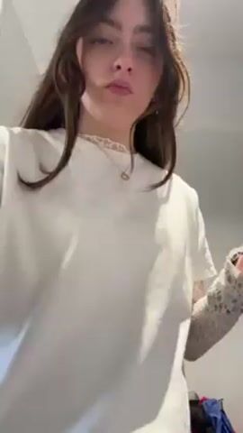 A young woman with long brown hair stands indoors and removes a pair of glasses, then pulls a white t-shirt over her head, pausing momentarily with the shirt obscuring her face before continuing to pull it off, revealing a long-sleeved lace undershirt.