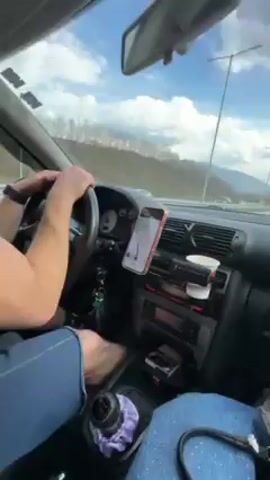 A man is driving a car on a highway. He has a beard and is wearing a black shirt and blue pants. The car has a phone mounted to the dashboard, displaying a navigation app.  There is a cup in the cupholder. Outside the window, there is a view of mountains and trees under a cloudy sky. The road appears to be somewhat busy with other cars.