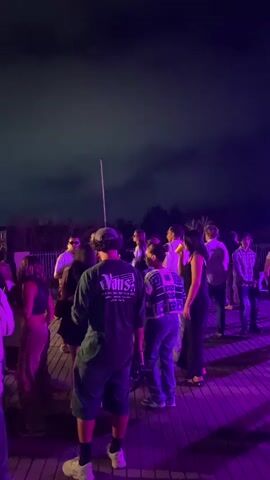 The video shows a nighttime outdoor gathering with a group of people dancing. The scene is illuminated by purple and yellow lights, suggesting a party or event. In the background, a DJ booth with equipment and lights is visible. The crowd appears to be a mix of men and women, dressed in casual attire. The ground is wooden, resembling a deck or patio. The sky is dark, indicating it is night.