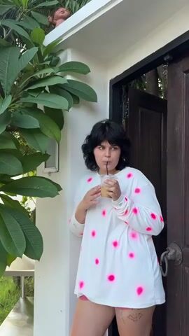 A woman with dark, curly hair stands outside a doorway holding a cup of iced coffee and a straw. She is wearing a long-sleeved white shirt with bright pink spots and appears to be wearing shorts. A large green plant with broad leaves is to her left. The doorway behind her is made of dark wood. There is a small, light-colored object on the ledge above the doorway. The setting appears to be outdoors.