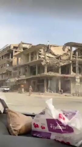The video shows a street view from inside a vehicle, with the camera pointed towards a heavily damaged building across the street. The building appears to be multi-storied, with significant structural damage, including missing walls and ceilings. There are tires stacked next to the building. Pedestrians, including a woman in traditional Middle Eastern clothing, are walking near the building. A motorcycle with two riders passes by on the street. Inside the car, a bag of what appears to be bread and a package of tissues are visible. The sky is overcast.