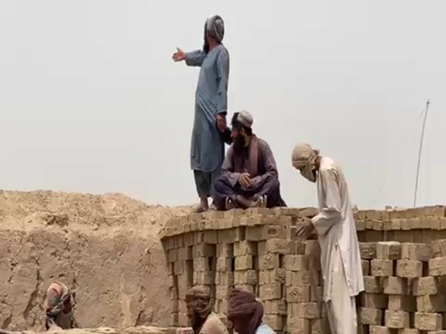 Several men are gathered on and around a stack of bricks. One man stands on the bricks, gesturing with his right hand, while another man sits cross-legged next to him. A third man in a white robe and face covering is placing bricks. Two other men with covered faces can be seen at the bottom of the stack. The environment appears to be a dusty, outdoor work site with a bright, overcast sky.