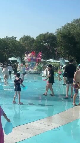 The video shows a sunny day at a water park. There are many people, including children and adults, playing in shallow water with fountains and water features. Some people are holding buckets and water guns, while others are simply enjoying the water. In the background, there are trees, buildings, and a tall structure with a ride. The water is clear blue, and the sky is bright. There are also inflatable toys visible in the water.