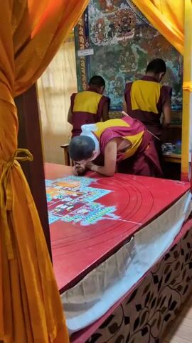 Here is a detailed description of the video:

The video shows three monks working on a sand mandala. One monk is bent over a low red table, carefully adding sand to the mandala with a stylus-like tool. The table is draped with a white cloth with a floral pattern. Another monk stands to the left, and another stands to the right of the table, both wearing similar maroon and yellow robes, appearing to assist. A tapestry with detailed images and figures hangs on the wall behind them. A yellow curtain is draped in the foreground, partly obscuring the view. The light is soft and warm, highlighting the intricate details of the sand mandala being created.