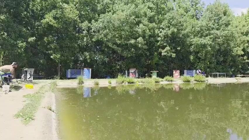 The video shows a body of water, likely a pond or lake, with a tree-lined bank. Along the bank, there are several small structures, some of which appear to be booths or shelters, and some people are visible, possibly fishing. The water is green and reflects the trees and sky. The overall scene suggests an outdoor recreational area on a sunny day.