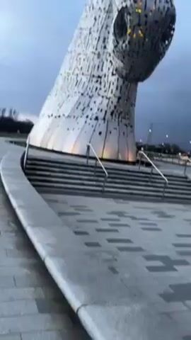 The video shows a low-angle pan up to reveal two large horse head sculptures made of silver-colored metal panels. The sculptures are set against a dark, cloudy sky. There are concrete steps leading up to the sculptures, with metal railings on either side. The ground around the sculptures is paved with gray and black patterned stones. There are streetlights in the background.
