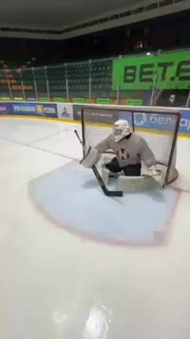 A hockey goalie wearing full protective gear stands in front of a net on a white ice rink. The goalie is facing the camera, then turns to their right as a puck is shot at them. The goalie drops to a knee to block the puck. In the background are rows of empty green seats and advertising banners on the walls.
