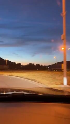 The video shows a view from inside a vehicle at dusk. The sky is a deep blue with scattered clouds, illuminated by the last rays of sunlight near the horizon. Streetlights and vehicle headlights cast a warm, yellow glow on the surroundings.

In the foreground, the dashboard and part of the windshield of the vehicle are visible. Outside the vehicle, a wide, flat landscape stretches out, with a road or highway running through it. In the distance, there are hills or mountains, silhouetted against the colorful sky.

Various objects are scattered across the landscape, including what appear to be buildings or structures, and a long train or series of train cars. A tall streetlight stands prominently on the left side of the frame. Trees with autumnal foliage are also visible, particularly on the right side of the frame, where a covered seating area with red accents can be seen. The overall impression is of a rural or semi-rural setting during twilight.