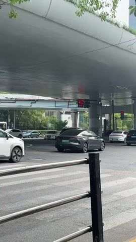 The video shows a street scene in China. A raised highway overpass dominates the background, casting shadows on the street below. Several cars, including a white SUV, a dark sedan, and a gray hatchback, wait at a crosswalk under a traffic light. A yellow shared bicycle is parked on the sidewalk next to a tree with green leaves. The street is paved with concrete, and the sidewalks are tiled. The sky is overcast.