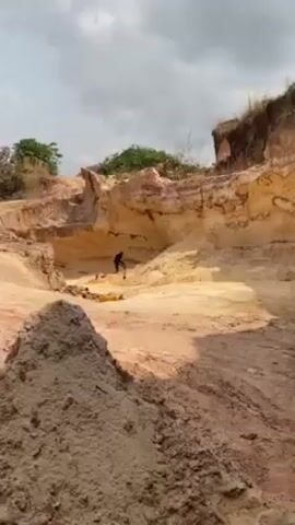 The video shows an adult person digging in a quarry. The person is in the middle distance and is wearing dark clothing. The person is digging into a light-colored sandy wall of the quarry with a pickaxe. The quarry is surrounded by hills of similar material. There is some sparse vegetation visible on the top of the hills. The sky is overcast.
