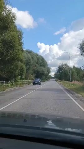 A black SUV is driving on a two-lane road. The road is surrounded by trees on both sides. The sky is blue with some white clouds. The weather appears to be sunny and warm. The SUV is in the left lane, moving away from the camera. The camera is positioned inside another vehicle, looking forward through the windshield. The road has white lane markings. There is a metal fence on the left side of the road. A lamppost is visible on the right side of the road. The terrain is flat.