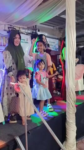 A woman in a green hijab and patterned dress stands on the left of a brightly lit stage, holding the hands of two young children. The youngest child, a toddler in a beige dress, stands to her left. A girl, approximately five years old, in a light blue denim overall dress and pink top, stands to her right, smiling and looking towards the right. Behind the young girl, an older child, approximately ten years old, in a light t-shirt with text, stands with one arm raised. To the right, a woman in a dark red off-the-shoulder dress sings into a microphone. Another person in a dark purple dress is partially visible behind the singer on the far right. The stage features colorful lights and is adorned with white draped fabric. Speakers are visible on the stage.