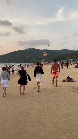 The video shows a beach scene at dusk. The sand is light brown, and a crowd of people is visible, strolling along the beach and enjoying the water. A hill covered in green vegetation serves as a backdrop. Above the water, a parasail is airborne. People in the foreground include a woman in a black dress carrying a blue jacket, another woman in a white skirt and black top, and a man wearing orange shorts. The sky is overcast with the sun setting, casting a warm glow.