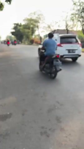 The media file captures a street scene in Kludan, East Java. The time of day appears to be late afternoon, with long shadows cast by the trees lining the road and the warm, soft sunlight indicating favorable weather.

The primary subjects are people and vehicles. On the road, a white SUV with the license plate "B 1970 HAM" is traveling away from the camera. Two individuals are seen riding motorcycles. One is on a black scooter directly in front of the SUV, and another is on a lighter-colored motorcycle further to the right, moving in the same direction. Several other motorcycles are visible in the background, suggesting moderate traffic. The road itself is paved and appears to be in good condition.

The setting includes a tree-lined street with mature trees providing shade. Beyond the trees, residential buildings with light-colored walls and tiled roofs are visible, characteristic of a semi-urban or suburban environment. The overall atmosphere appears calm and ordinary, depicting a typical day on a local road. There are no overt signs of distress or excitement among the people, suggesting a routine street scene. The lighting and the presence of foliage indicate a pleasant day.