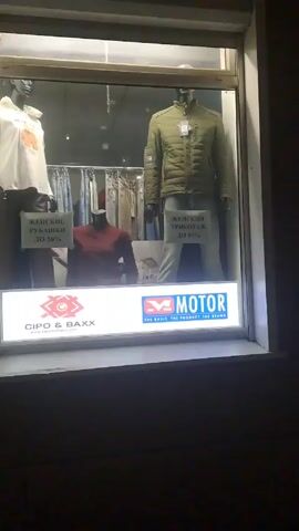 The video shows a series of store windows displaying clothing. The first window features mannequins wearing jackets and shirts with signs advertising a sale. The window also shows the logos for the clothing brands Cipo & Baxx and Motor. The second window showcases Wrangler and Lee clothing brands, featuring mannequins in vests, shirts, and jeans. The third window, similar to the second, displays Wrangler and Lee clothing on mannequins. The video is taken at night and the windows are illuminated.