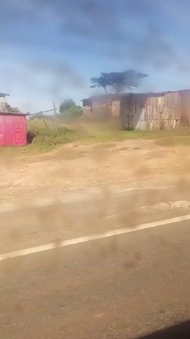 The video shows a landscape shot from a moving vehicle on a clear, sunny day. The road is paved and has a solid white line dividing the lanes. On the right side of the road is a dirt area with several structures that appear to be small, informal businesses or dwellings made of corrugated metal. One of these structures has a sign that reads "4G UPGRADE TO THE FAST LANE." 

Several vehicles are parked in the dirt area, including a white sedan, a black SUV, and a yellow school bus. Further along, there is also a blue bus. People are seen standing and walking around the vehicles and structures. In the background, a distant mountain range is visible. A tall street light stands near the edge of the dirt area. The overall impression is of a roadside scene in a rural or developing area.