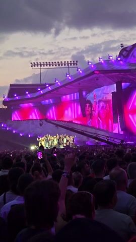 This is a vertical video captured during an outdoor music concert in Cluj-Napoca, Romania. The scene is dominated by a large, elaborate stage illuminated by vibrant pink and purple lights, with a large screen displaying imagery. A crowd of people is gathered in front of the stage, facing towards it, with many holding up their phones to record or take pictures. The time of day appears to be late afternoon or early evening, as indicated by the soft, cloudy sky. The overall atmosphere is one of excitement and engagement, typical of a live music event. There is no visible text in the video.
