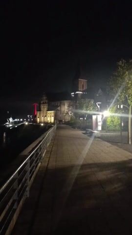 The video shows a nighttime scene along a waterfront promenade. A metal railing runs along the left side of the frame, separating the walkway from the dark water. In the distance, a large building with a tall tower is illuminated with yellow lights and a single red light. To the left of the building, more lights can be seen along the water's edge. A street lamp illuminates the promenade on the right side of the frame, casting long shadows. There are trees to the right of the promenade. The sky is completely dark.