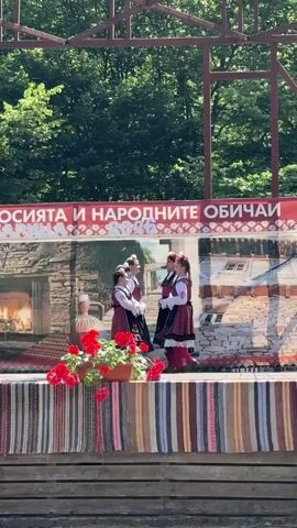 A group of six young women wearing traditional Bulgarian folk costumes perform on an outdoor stage. The costumes consist of white blouses, dark vests with red trim, and layered skirts with intricate embroidered patterns in red and black. They also wear red headbands with decorative elements and dark shoes. The women are arranged in two rows, with four in the back and two in the front. They are holding hands in a line formation and appear to be dancing or posing. In the foreground, a brown planter filled with bright red geraniums sits on the edge of the stage. The stage is decorated with a colorful, striped fabric runner. Behind the performers, a large banner with Cyrillic text and a backdrop depicting a traditional village scene are visible. The backdrop features stone buildings, a fireplace, and hints of nature. The stage is set against a natural background of lush green trees and foliage, with a glimpse of a metal structure above, suggesting an open-air venue or festival. The lighting indicates it is daytime.