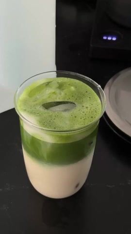 Here is a description of the video.

A person uses a clear straw to stir an iced matcha latte in a clear glass on a black countertop. The drink has layers of white milk at the bottom, a layer of green matcha, and a frothy layer of green foam at the top. There is an ice cube in the center of the foam. A white water filter and a stainless-steel electric tea kettle are in the background.