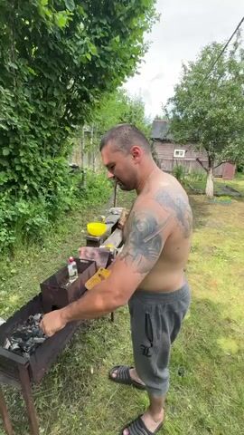 A shirtless man with a tattoo on his back and arm is standing in a grassy yard preparing a barbecue. He is wearing grey sweatpants and black sandals. He is using a metal poker to arrange coals in a dark metal barbecue grill. Next to the grill, there is a yellow bowl, a bottle of liquid, and a small white bag. In the background, there are lush green trees and a wooden house with a white window. The sky is overcast. The man appears to be focused on his task.