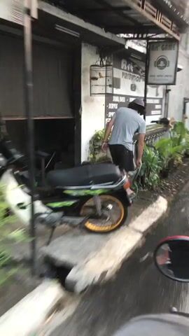 A person wearing a grey t-shirt, black shorts, and a grey baseball cap is leaning over and appears to be watering plants with a green hose. They are standing next to a parked motorcycle which is partially visible in the foreground and mid-ground. The motorcycle has a green and black seat and a yellow rim on its rear wheel. The person is facing away from the camera. The background shows a building with a sign that reads "GAME". There are also plants and a birdcage visible. The ground appears wet, suggesting recent watering or rain. The scene is outdoors, possibly a street or alleyway. The image is in motion blur, indicating the video is taken from a moving vehicle.
