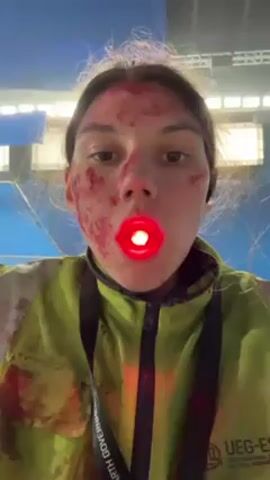A young woman films herself from the chest up. Her face and jacket are spattered with red, likely blood or a similar substance. Her hair is messy. There is a small red light inside her mouth, illuminating it from within. She wears a bright yellow jacket with writing on the left chest, including the words “EARTH GOVERNMENT”. She also wears wireless earbuds. The background is blurred, appearing to be inside a large indoor structure with a blue ceiling and overhead lights.