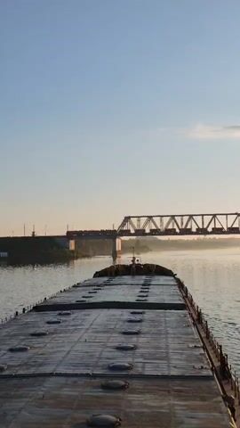 The video shows a barge navigating a river beneath a railway bridge. The barge has a flat, dark-colored deck with several circular access points.  A train is crossing the bridge, which is a steel truss structure supported by concrete piers. The river is calm, and the sky is clear with a slight haze near the horizon. The banks of the river are lined with trees and greenery.  The light suggests it is either early morning or late afternoon.