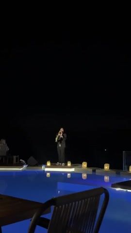 A person in a suit stands on a raised platform in front of a pool at night, playing a saxophone. Several small, lit lanterns are placed around the platform. In the background, two people sit at a table. The sky is dark, with some faint lights visible. A portion of a table and chair is in the foreground.