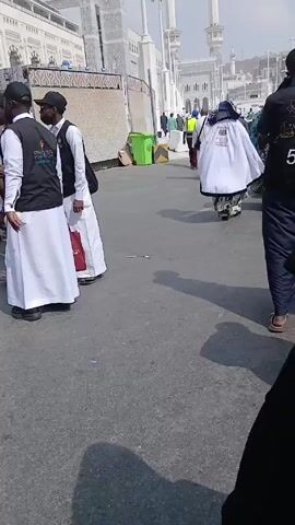 The video shows people walking on a paved surface near a large, white building. Some individuals are wearing white robes, while others are dressed in black and white attire, including vests. A police officer is also visible. A green trash bin and construction barriers are present in the background. The building has a symmetrical design with arches and towers.
