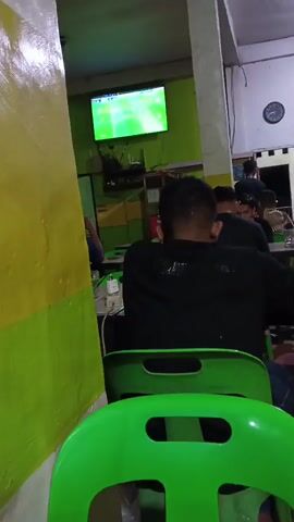The video shows the interior of a dimly lit establishment with people seated at tables, facing a television screen displaying a soccer match. The camera is positioned behind a person wearing a black hooded sweatshirt, their back to the camera. Green plastic chairs are visible in the foreground. The walls are painted in shades of yellow and green. In the background, other individuals are seen seated and interacting. A shelf unit is visible with various items on display. A clock hangs on the wall. The overall atmosphere appears casual, with patrons focused on the television.