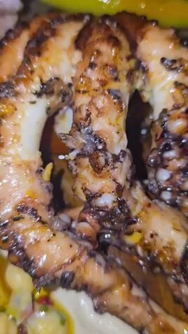 A close-up shot of a grilled octopus dish is presented on a wooden serving board. The octopus, charred in places, rests on a smooth bed of hummus. Chickpeas and a green herb garnish with a drizzle of oil are visible on the hummus next to the octopus. As the camera slowly zooms out, more elements of the dish become visible: crispy yellow chips or crackers, potato wedges, and a green pepper. A purple-colored, ruffled item is also on the board. The setting appears to be an indoor dining environment with warm lighting. Soft background music is audible throughout the video.