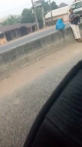 The video shows a view from inside a vehicle, looking out across a street and a concrete barrier. On the opposite side of the street, a woman stands on the edge of the concrete barrier, next to a blue bag. The woman is wearing a patterned top and light-colored pants. In the background, there are various buildings and trees, and utility poles are visible. A vehicle can be seen passing by on the street. The asphalt road in the foreground shows wear and tear. The weather appears overcast.