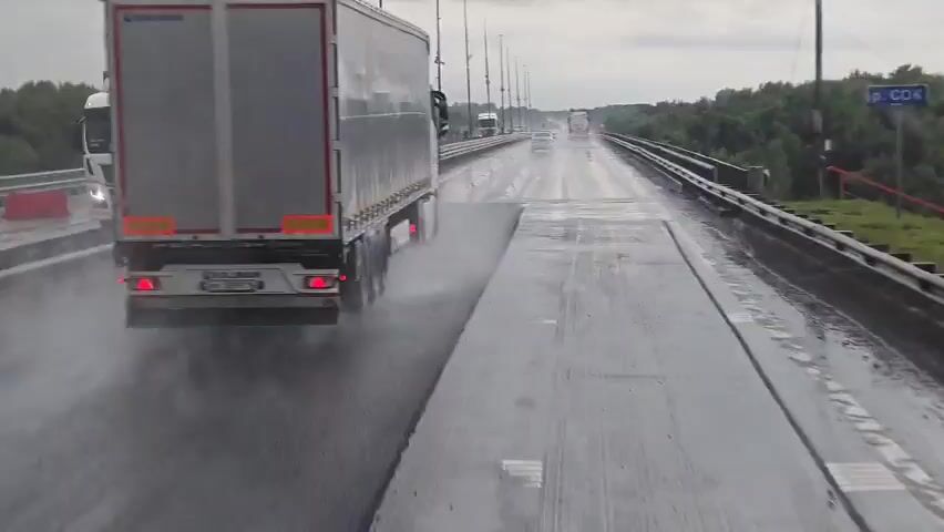 The video shows a wet highway on a cloudy day. The camera is looking forward from a vehicle, likely a truck given the view from the cab. A large white truck with a silver trailer is in front and to the left, spraying water as it drives. Several other vehicles are visible further down the highway, also driving in wet conditions. A metal guardrail runs along the right side of the road, with trees and dense foliage visible beyond it. The road surface appears to be asphalt and is reflecting the grey sky. The weather appears to be rainy or recently rained.