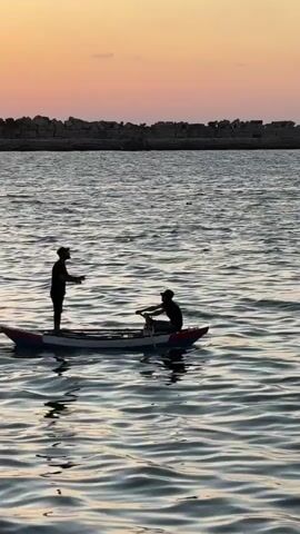 Two silhouetted figures are in a small boat on the water. One person is standing at the front of the boat, facing the other person, and appears to be gesturing with their hands. The second person is seated at the back of the boat, holding oars and appears to be rowing. The water is choppy, with visible ripples. In the background, there is a breakwater made of large rocks, and a hazy sky with a gradient of colors suggesting either dawn or dusk. The overall lighting is dim.