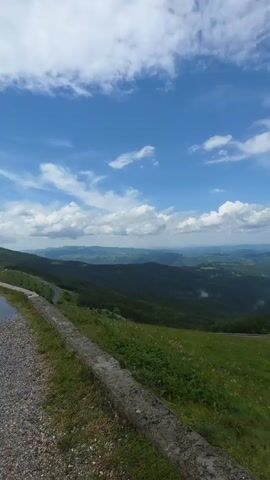 The video opens with a panoramic view of rolling green hills under a partly cloudy sky. A winding road is visible in the distance, snaking through the landscape. The camera pans to the right, revealing more of the lush terrain and a glimpse of a distant town. As the video progresses, the viewpoint shifts to a gravel road with a stone retaining wall on the left. A dark SUV is parked on the right side of the road. The camera then moves closer to a large, arched monument made of concrete. Two stone figures flank the entrance to the arch, and the year 1876 is inscribed above it. Several cars are parked near the monument. The sky is filled with dramatic, heavy clouds.
