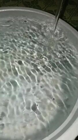 A close-up shot shows water being poured from an unseen source into a round, silver basin filled with water. The water in the basin is disturbed by the pouring, creating ripples and waves that spread across the surface. The lighting is dim, and the background is out of focus.