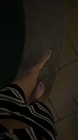 A low-angle shot starts focused on a person’s foot with white nail polish on a ledge next to a pool, then pans out and up. The person is wearing a striped skirt. The pool water is dark. The view then rises to reveal a rooftop pool area at night with patio furniture (lounge chairs, small tables) and plants.  In the background, there is a cityscape with tall buildings lit up. The night sky is dark.
