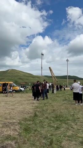 A group of people are gathered in a grassy field with rolling hills in the background under a cloudy sky. In the center of the frame, a tall wooden pole stands upright, with two individuals climbing it. One person is near the top of the pole, attached by a rope, while another is further down. To the right of the pole, a large yellow crane-like structure is present. A yellow van is visible on the left side of the frame, with several people standing near it. Other individuals are scattered throughout the field, observing the activity. The overall scene appears to be an outdoor event or competition.