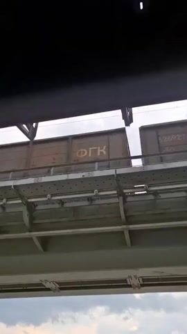The video shows a view from below a bridge, looking up at a train passing overhead. The train consists of several open-top freight cars, likely carrying some kind of cargo. The cars are dark in color, possibly green or gray, and have various markings and numbers visible. The train is moving from left to right across the frame. The bridge itself appears to be made of concrete and steel, with visible structural supports and walkways. The sky is overcast with clouds visible. The lighting suggests it is daytime.