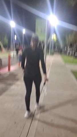 A woman with fair skin and pulled-back hair is walking on a sidewalk next to a park at night. She is wearing a black long-sleeved shirt, black leggings, and white sneakers. She is carrying a cell phone in one hand and a plastic water bottle in the other. As she walks, she turns the camera to face herself and talks, expressing surprise or frustration. The park features playground equipment, picnic tables, and groups of people. Streetlights illuminate the scene.