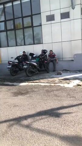 Two men are sitting on a ledge next to two motorcycles. The man on the left is wearing a dark jacket and a beanie. The man in the middle is wearing a grey shirt and dark pants. The man on the right is wearing a striped shirt and dark pants. The motorcycles are parked on a paved surface. The background appears to be a building with large windows.